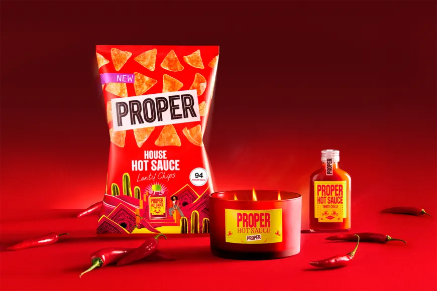 PROPER House Hot Sauce Candle lit beside a packet of House Hot Sauce lentil chips and a bottle of PROPER hot sauce on a red background