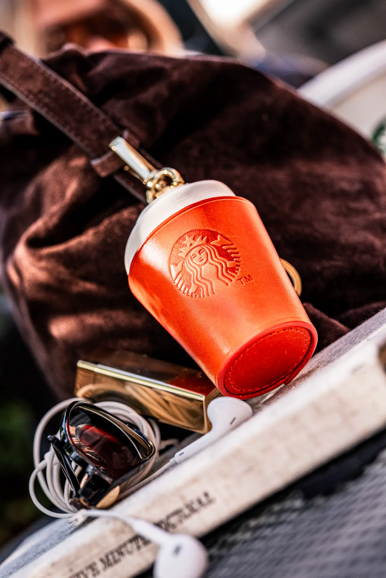The Autumn Charm leather coffee-cup bag charm with gold clasp clipped to a brown suede handbag beside sunglasses and earphones, made by The Robin Collective